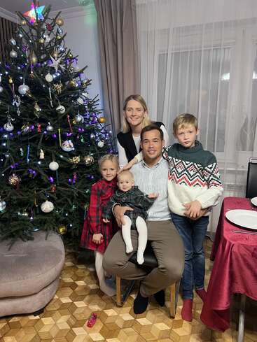 A happy family of five poses near a decorated Christmas tree. The parents and three children smile warmly, celebrating together in a festive, cozy living room.