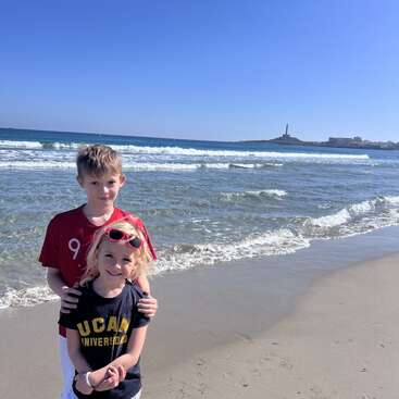 Two children stand together on a sunny beach, smiling. Gentle waves roll in behind them, with a distant lighthouse visible under a clear blue sky.