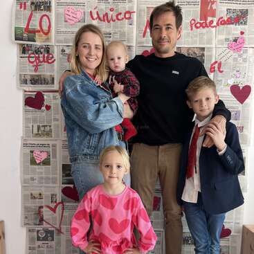 A smiling family of five poses in front of a newspaper backdrop with red hearts and Spanish writing, dressed casually and semi-formally, radiating warmth and togetherness.