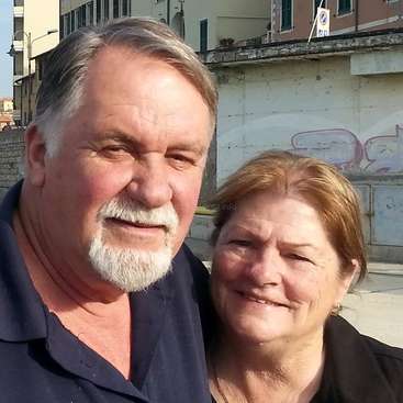 The image shows an older couple posing for a photo in front of a city street with buildings and graffiti, likely taken on a sunny day.