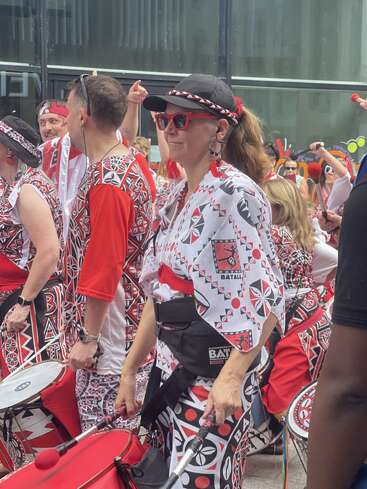 Un groupe de tambours dynamiques, vêtus de tenues à motifs rouges, noirs et blancs assortis, se produisant en plein air. Une femme portant des lunettes de soleil et un chapeau dirige le groupe avec enthousiasme, tambours visibles.