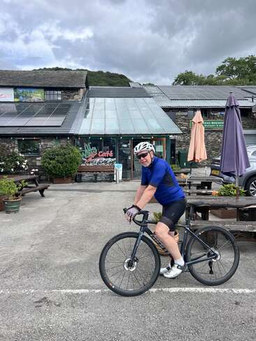 Un cycliste en tenue bleue et casque pose avec son vélo devant un café rustique avec des panneaux solaires, des bancs, des plantes en pot et un ciel nuageux au-dessus de sa tête.