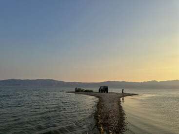 A narrow, rocky path stretches into a serene lake at sunset. A person and a vehicle stand at the end, mountains silhouetted in the background.