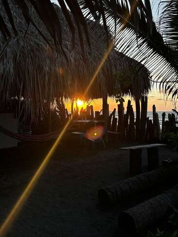 La imagen representa una serena escena playera al atardecer, con una cabaña de paja, palmeras y una hamaca, y el sol poniéndose sobre el océano al fondo.