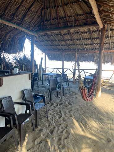 Esta imagen muestra una cabaña de playa rústica con techo de paja, sillas de plástico, una hamaca, suelo de arena, bar de madera y un ambiente tropical relajado y acogedor junto al mar.