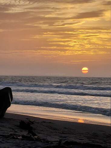 Una puesta de sol dorada ilumina suavemente las olas del océano, con nubes dispersas en el cielo y una playa tranquila y vacía en primer plano, que arroja tranquilos reflejos.