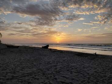 Una tranquila escena de playa al atardecer, con barcas descansando en la arena, suaves olas, nubes dispersas y un cálido sol dorado que ilumina bellamente el sereno horizonte.
