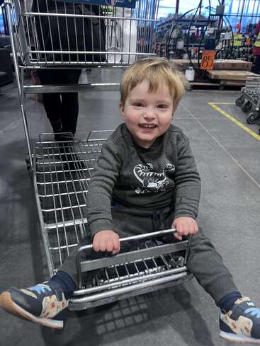 Um menino alegre senta-se brincando em um carrinho de compras de metal em uma loja, sorrindo brilhantemente. Ele usa uma roupa cinza e tênis coloridos.