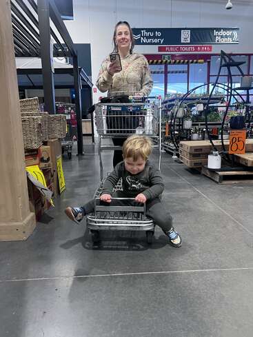 Uma mulher sorridente empurra um carrinho de compras em uma loja, enquanto uma criança pequena senta-se brincando em um carrinho pequeno, aproveitando a aventura de compras juntos.
