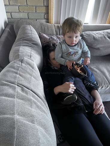 Uma criança pequena senta-se no colo de uma mulher em um sofá cinza, comendo de uma tigela. A mulher está deitada embaixo dele, olhando para cima; um momento aconchegante e casual.