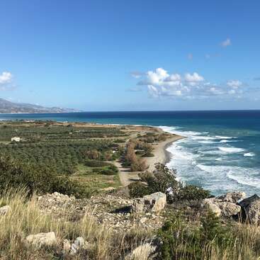 Un hermoso paisaje costero con una costa rocosa, olas ondulantes, campos verdes, casas dispersas y montañas lejanas bajo un cielo azul claro con nubes esponjosas.