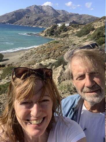 Una pareja sonriente se hace un selfie cerca de una costa pintoresca. Detrás de ellos hay montañas escarpadas, mar azul, playa de arena, arbustos dispersos y algunas casas blancas.