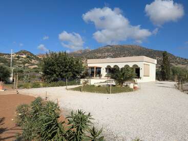 Una casa blanca con arcos se encuentra en un paisaje rural, rodeada de vegetación, un camino de grava, montañas lejanas y un cielo azul brillante con nubes.