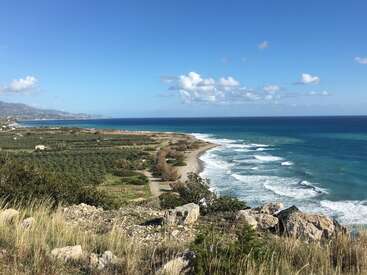 Un magnifique paysage côtier avec un rivage rocheux, des vagues qui roulent, des champs verts, des maisons éparpillées et des montagnes lointaines sous un ciel bleu clair avec des nuages floconneux.