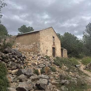 Une vieille maison en pierre abandonnée se dresse dans une zone rurale, entourée de gravats et de végétation sauvage. Le ciel nuageux ajoute une touche mélancolique et atmosphérique à la scène.