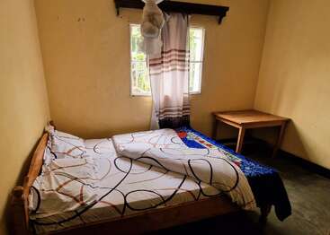 Ein bescheidenes Schlafzimmer mit gelben Wänden, einem Holzbett mit gemusterten Laken, einem Tisch in der Ecke, mit Vorhängen verhangenen Fenstern und einem hängenden Moskitonetz.