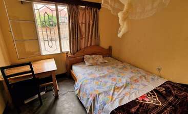 Dieses Bild zeigt ein kleines, gemütliches Schlafzimmer mit einem Holzbett, bunter Bettwäsche, einem Schreibtisch und Stuhl, einem offenen Fenster, Vorhängen und einem darüber hängenden Moskitonetz.