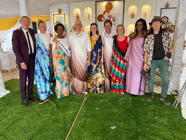 A group of people stands together, smiling, dressed in colorful traditional attire. They are inside a decorated venue with white walls, green grass, and festive ornaments.