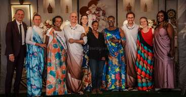 A diverse group of people stands together, smiling, dressed in colorful traditional African attire and formal wear, against a decorated background with flowers and art pieces.