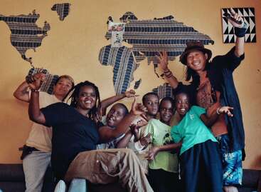 A joyful group of adults and children pose together, smiling and making fun gestures, in front of a decorative world map on a warmly colored wall.