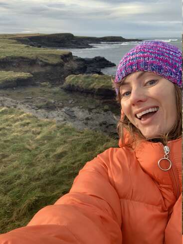 Una mujer sonriente, con una chaqueta naranja y un gorro de punto de colores, se hace un selfi cerca de una costa escarpada con acantilados cubiertos de hierba y el mar de fondo, bajo un cielo nublado.