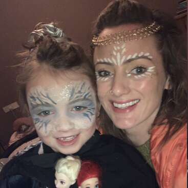 Una mujer y un niño, con la cara pintada de forma creativa, sonríen juntos. El niño sostiene dos muñecas, y ambos parecen ir disfrazados, disfrutando del momento.