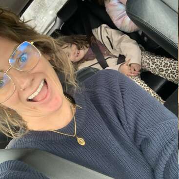 Eine fröhliche Frau mit Brille macht ein Selfie im Auto. Auf dem Rücksitz sitzen zwei Kinder und lächeln; eines trägt eine Hose mit Leopardenmuster und einen beigen Mantel.
