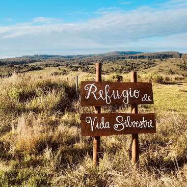 Un panneau en bois indique "Refugio de Vida Silvestre" dans un champ herbeux sous un ciel bleu vif, avec des collines et des forêts en arrière-plan. Sanctuaire de la nature.