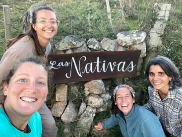 Four women are smiling happily and posing outdoors next to a rustic stone wall with a wooden sign that reads “Las Nativas” in beautiful cursive script.