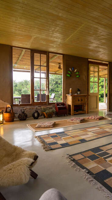A cozy rustic room with wooden ceiling, large windows, woven rugs, floor cushions, and lanterns. Outside, greenery and patio furniture create a peaceful, inviting atmosphere.