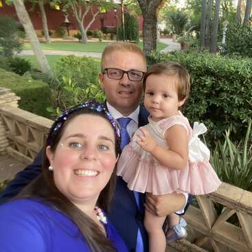 Das Bild zeigt eine dreiköpfige Familie, die für ein Selfie vor einem Garten posiert. Die Frau steht links, der Mann in der Mitte und das Kind rechts.