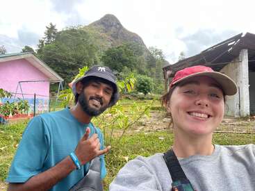 Um casal sorridente posa para uma selfie ao ar livre, perto de prédios coloridos e vegetação, com o pico de uma montanha se destacando ao fundo, sob um céu nublado.