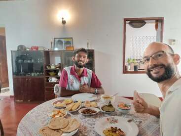 Dois homens estão sentados à mesa dentro de casa, sorrindo e fazendo um sinal de "joinha". A mesa está posta com uma variedade de deliciosos pratos indianos e pães achatados.