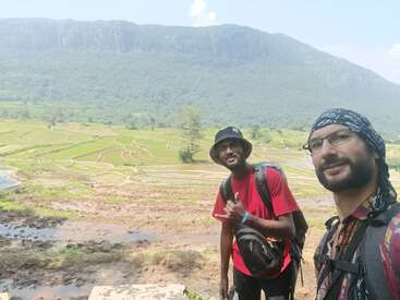 Dois homens tiram uma selfie em frente a campos em socalcos cobertos de vegetação exuberante e uma grande montanha, sob um céu azul brilhante, aproveitando uma aventura ao ar livre.