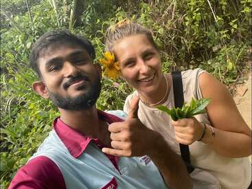 Um homem e uma mulher sorrindo ao ar livre, rodeados de vegetação. A mulher segura algumas folhas e tem uma flor amarela no cabelo. Eles parecem felizes, curtindo a natureza juntos.