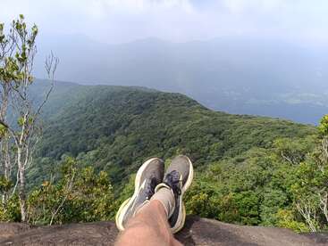 Uma pessoa relaxa à beira de um penhasco, com as pernas esticadas, contemplando um vasto vale verdejante e coberto de florestas, com montanhas enevoadas ao longe. Um clima tranquilo.