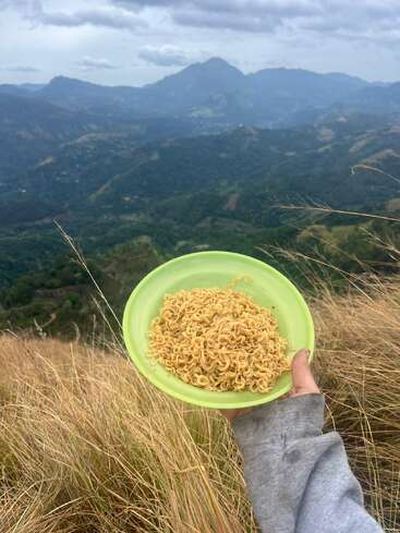 Em primeiro plano, uma mão segura um prato verde com macarrão cozido. Ao fundo, há grama alta, colinas onduladas e montanhas distantes sob um céu nublado.