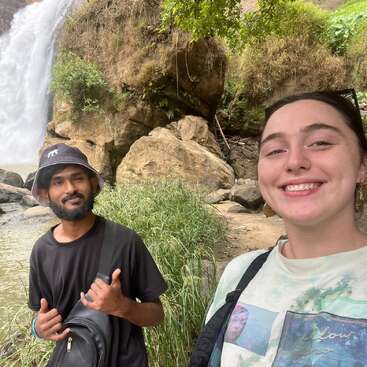 Um casal sorridente posa ao ar livre perto de uma cachoeira, cercado por rochas, vegetação e água, aproveitando uma caminhada pela natureza e tirando uma selfie.