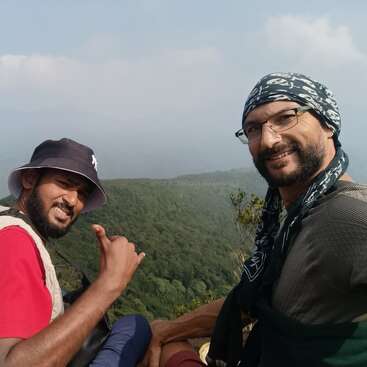 Dois homens estão sentados na encosta de uma montanha, sorrindo para a câmera. Eles vestem roupas casuais de caminhada e apreciam a vista panorâmica das colinas verdejantes sob um céu nublado.