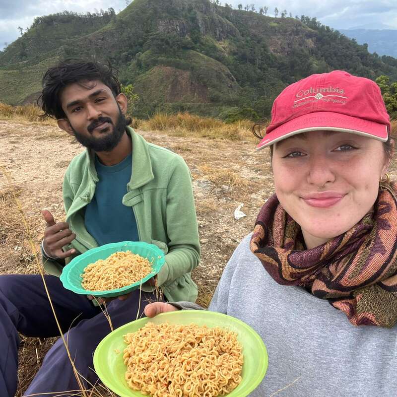 Um homem e uma mulher estão sentados ao ar livre com pratos de macarrão, sorrindo para a câmera. As montanhas e a vegetação criam um cenário natural e pitoresco para a refeição que compartilham.