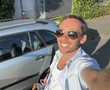 Un homme souriant portant des lunettes de soleil prend un selfie en plein air à côté d'une voiture argentée. Il est habillé de manière décontractée et semble heureux, profitant d'une journée ensoleillée.