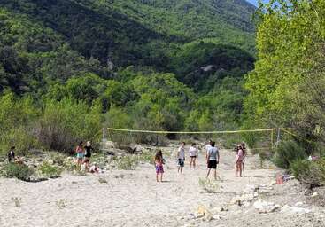 A group of people plays volleyball on a sandy, makeshift court surrounded by green trees and mountains, enjoying a sunny day outdoors in nature.