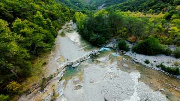 A tranquil river winds through a lush, green forest. A person stands on a stone bridge crossing the shallow waters, surrounded by rocky banks and trees.