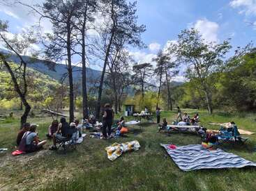 A group of people are having a picnic in a forested area, sitting on blankets and chairs, enjoying nature under a partly cloudy sky.
