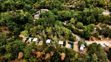 This aerial image shows a forested area with several tents or cabins nestled among the trees, winding dirt roads, and a few vehicles, creating a serene, secluded campsite.