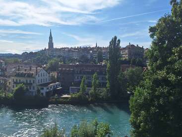 A picturesque European town with historic buildings, a tall church spire, lush greenery, and a vibrant blue river under a bright, partly cloudy sky. Peaceful scenery.