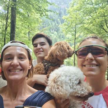 Three smiling people and two adorable poodles enjoy a beautiful sunny day outdoors, surrounded by lush green forest and mountains in the background, capturing a happy moment.