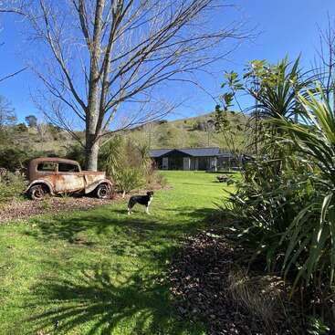 Das Bild zeigt eine ruhige ländliche Szene mit einem Oldtimer, einem Hund und einem Haus im Hintergrund, umgeben von üppigem Grün und einem klaren blauen Himmel.