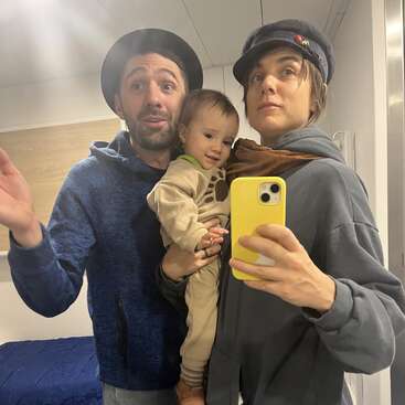 A family of three stands together for a mirror selfie. The adults wear hats and casual clothes, one holding a baby. Everyone looks cheerful and relaxed indoors.