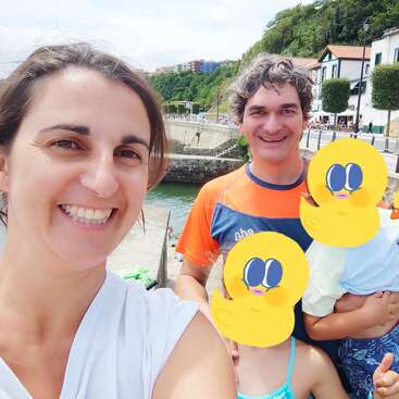 Das Bild zeigt eine vierköpfige Familie, die für ein Selfie auf einer Brücke über einen Fluss posiert, mit einer Stadt im Hintergrund und einem bewölkten Himmel.